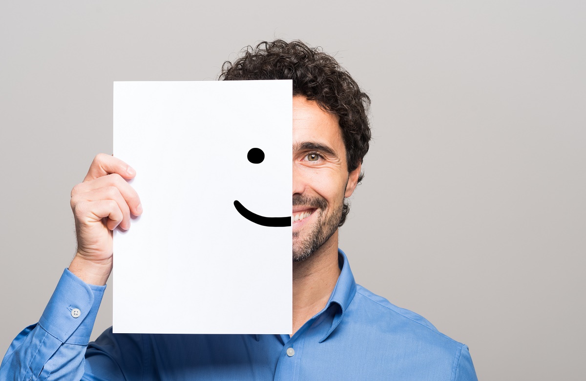 Man-holding-a-smiley-face-sign-on-one-side-of-face-while-smiling-TMS-for-Depression-how-it-helps-iMind-Mental-Health-Solutions-2