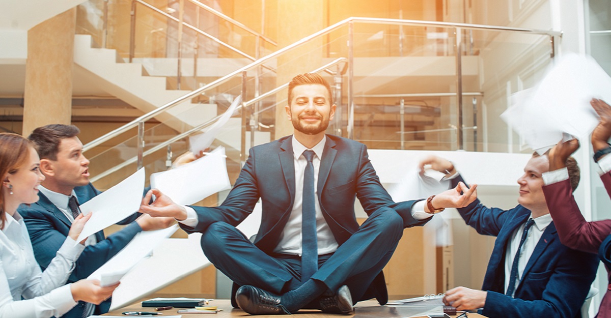 man-sitting-in-a-Zen-pose-on-a-conference-table-and-lit-from-above-while-coworkers-argue-11-ways-to-manage-workplace-stress-iMind-Mental-Health-Solutions