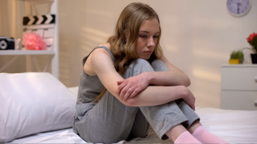 teenager-with-arms-crossed-over-legs-sitting-on-bed-and-looking-down-social-media-mental-health-teens-adolescents-iMind-Mental-Health-Solutions