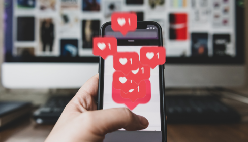 concept-of-social-media-hand-holding-phone-in-the-foreground-and-multiple-love-symbols-with-the-number-1-drawn-over-it-with-a-blurred-desk-and-computer-monitor-with-images-in-the-background-iM