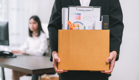 business-person-walking-toward-camera-holding-a-cardboard-box-of-belongings-while-colleague-in-background-looks-on-with-an-unhappy-look-on-her-face-how-to-spot-a-toxic-workplace-before-you-start.